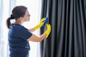 A woman wearing yellow rubber cleaning gloves is spot treating hanging grey curtains.