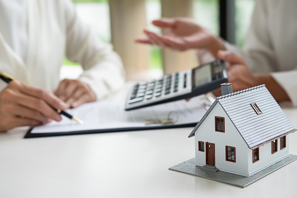 A small white model house sits on a table while two people discuss a document and numbers on a calculator.