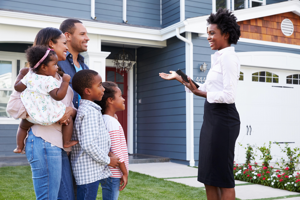 A woman in office wear speaks to a family of five in front of a house.