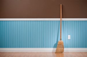 A wooden broom is standing on hardwood flooring and is leaned against a blue and brown wall.