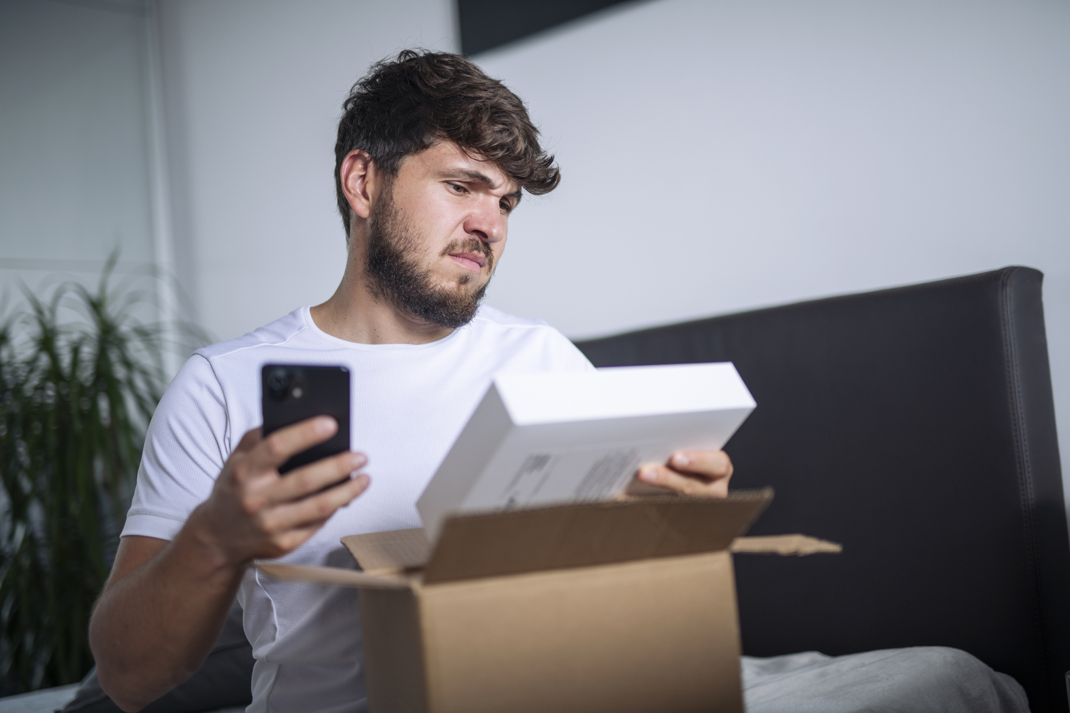 A young man looks confused at the product that was shipped to him, seemingly comparing it to the expected item on his phone.