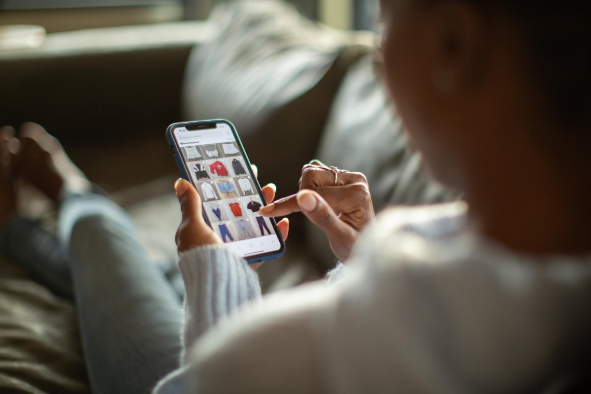 An out-of-focus woman is reclining on a sofa and scrolling through an online shopping app on her smartphone.