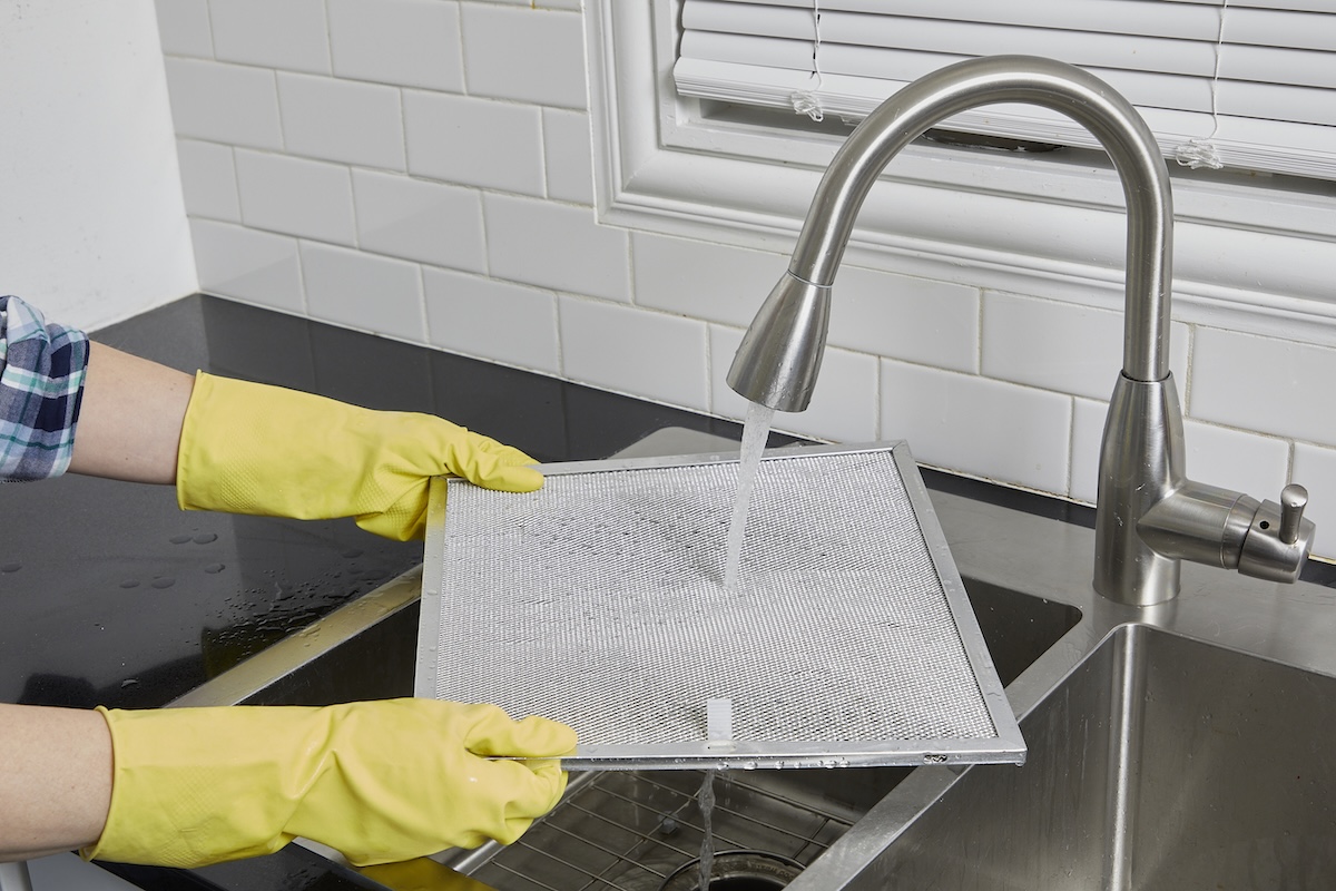 Woman wearing rubber gloves rinses a range hood filter under a kitchen faucet.