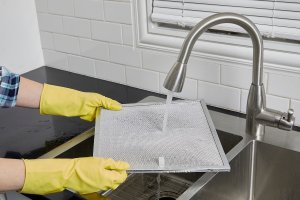 Woman wearing rubber gloves rinses a range hood filter under a kitchen faucet.