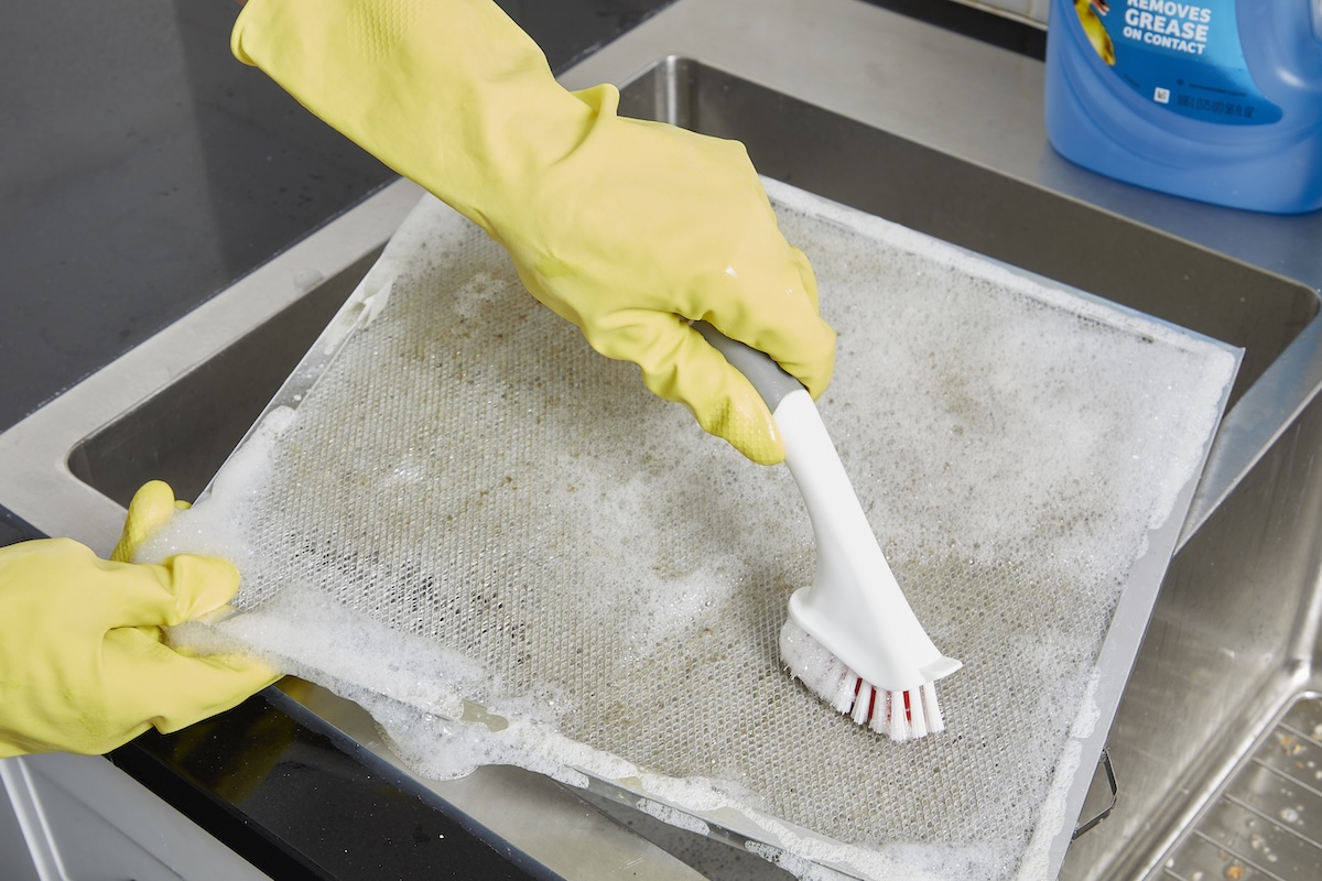 Woman wearing rubber gloves scrubs a range hood filter with a scrub brush and soapy water.