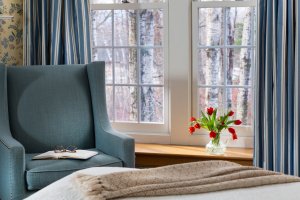 A spare bedroom window with open blue-and-white striped curtains, a blue reading chair, and made bed.