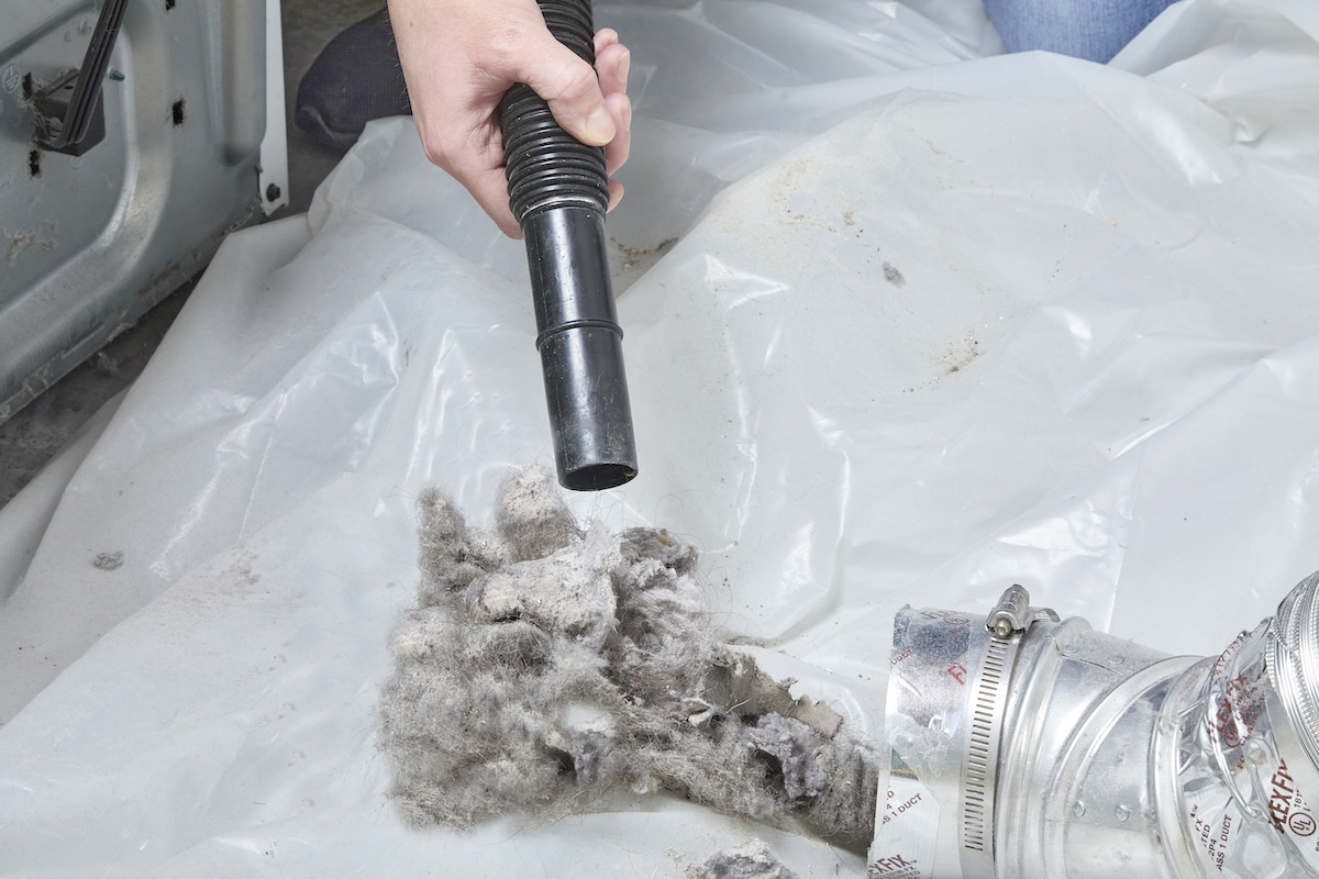 Person uses a vacuum attachment to suction lint off the floor after it is pulled out of a dryer vent.