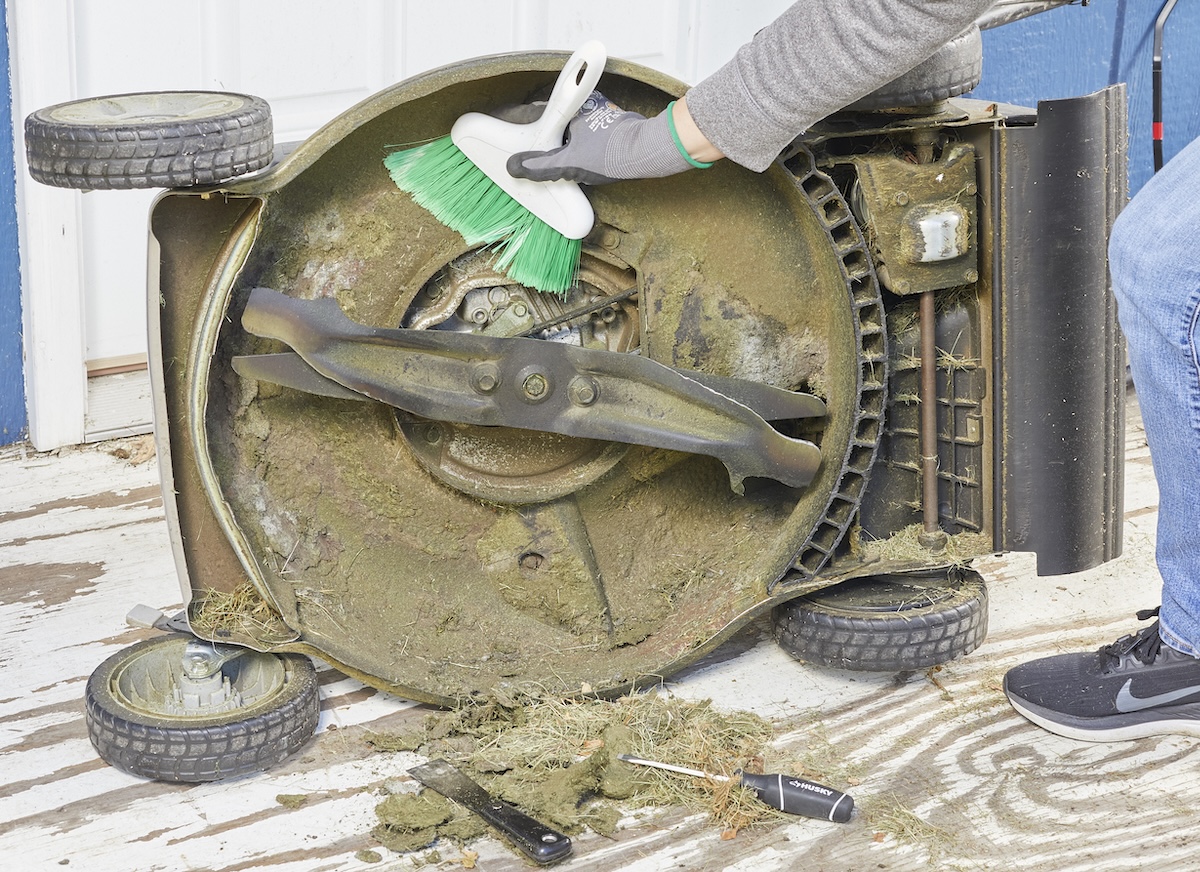 Woman cleans the underside of a lawn mower with a hand broom.