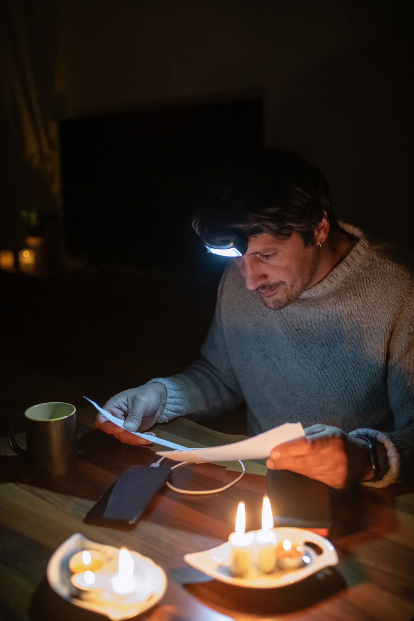 A seated man wearing a sweater and headlamp reads documents by candlelight during a winter power outage.