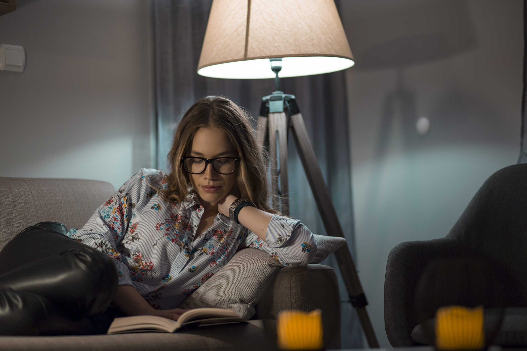 Young woman reading book in living room with lamp on in background.