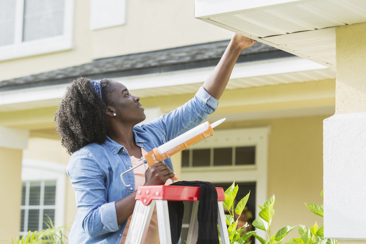 A homeowner on a ladder caulking gutters on a house.