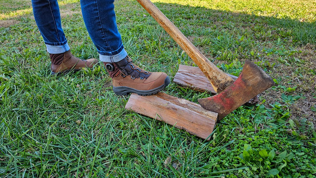 Person in jeans and brown red wing work boots stepping on fire wood and holding an ax