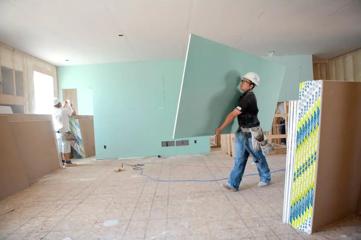 A DIYer or contractor carrying a sheet of green board drywall through a kitchen renovation site.