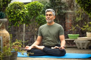 Man meditating in lotus position while sitting on exercise mat surrounded with plants in his backyard.