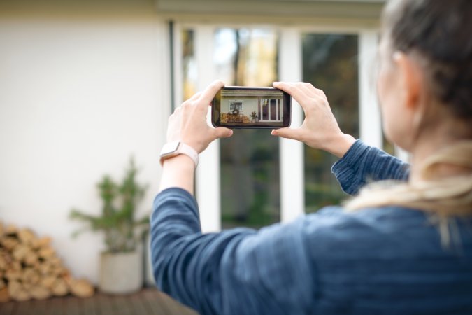 Older woman taking pictures of the outside of her house on a smartphone.