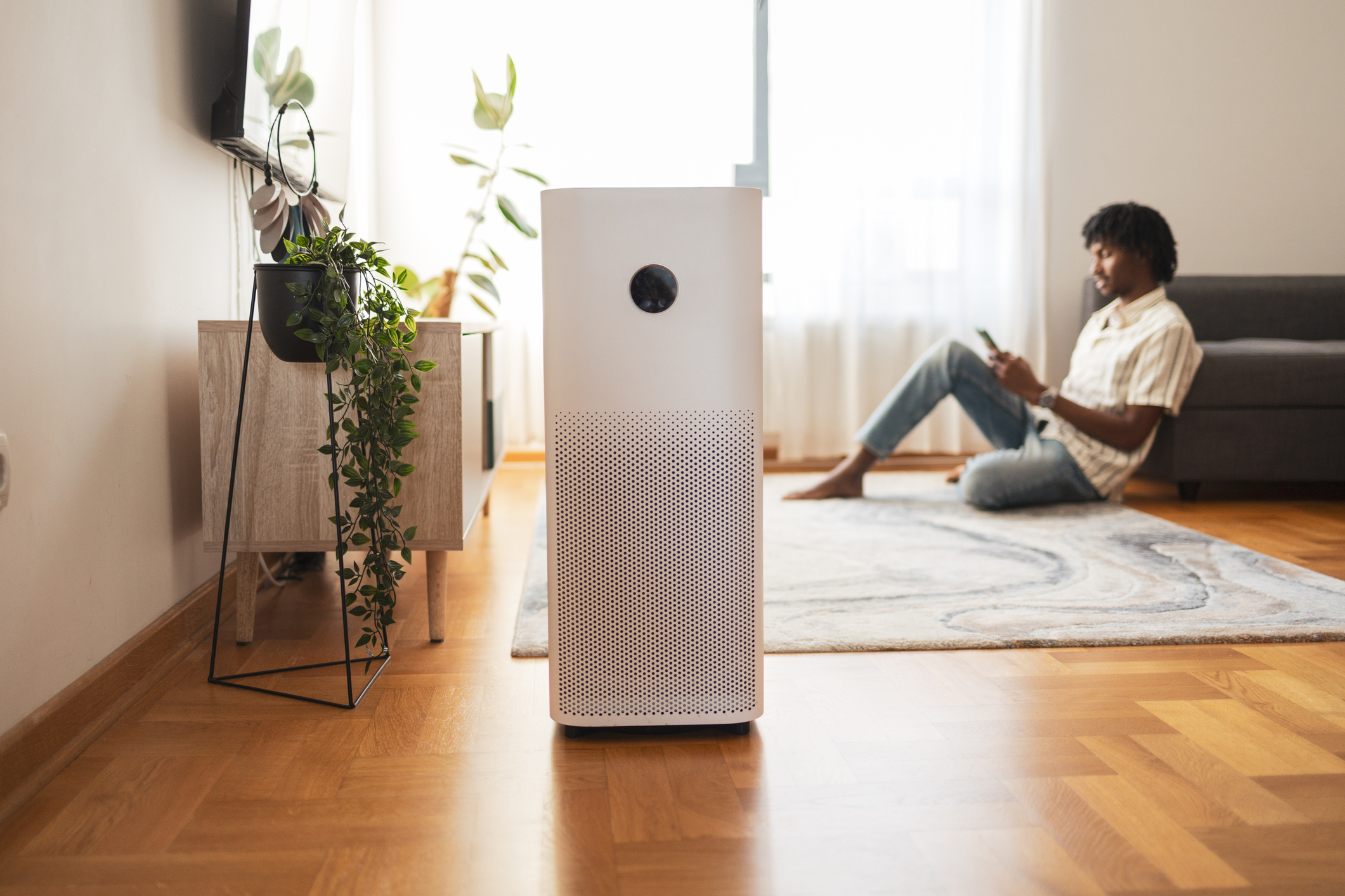 A man sitting on the floor across the room from an air purifier sitting on a wooden floor in a contemporary living room.