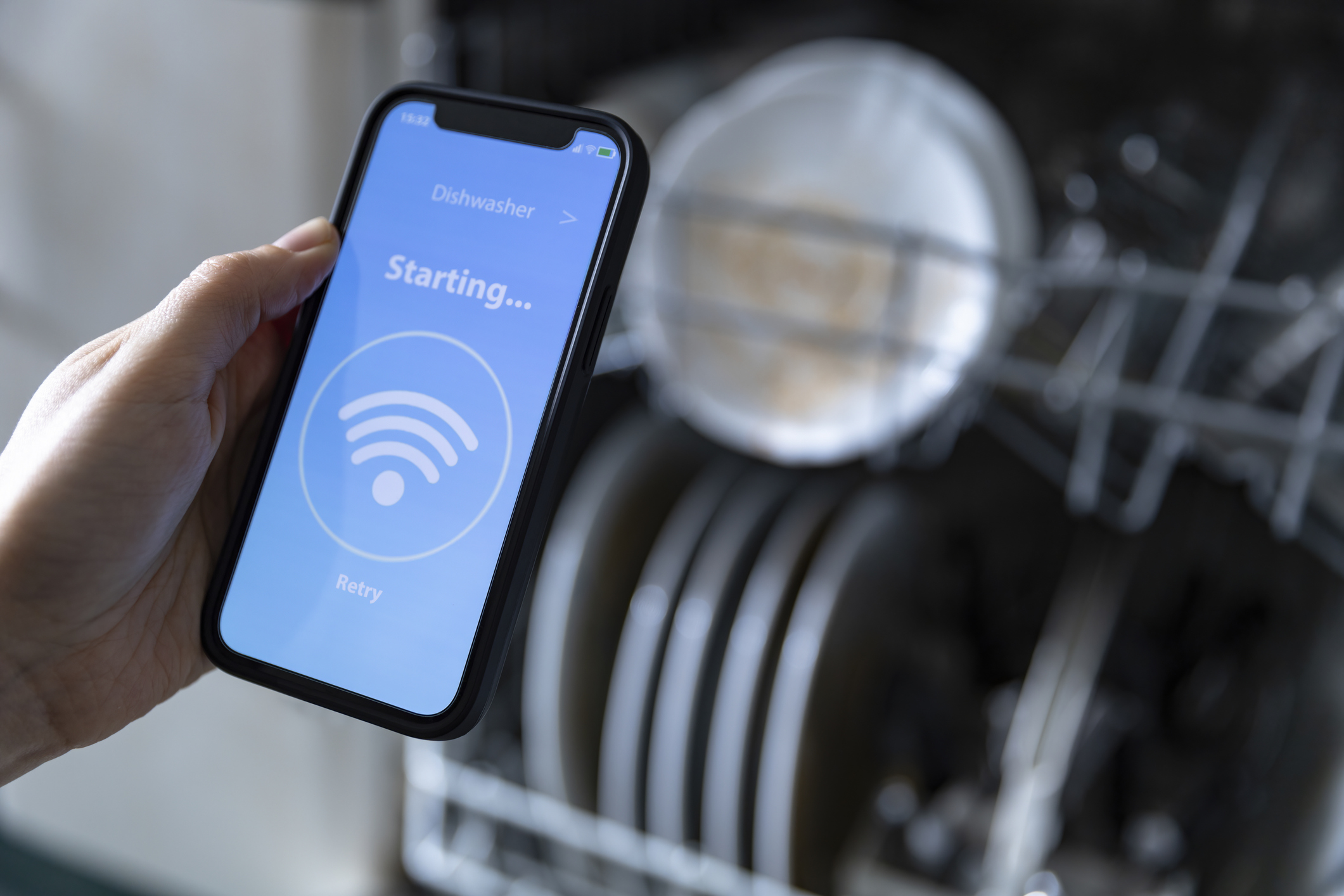 Hand holding a mobile phone app with a smart dishwasher app on the screen in front of a loaded dishwasher.