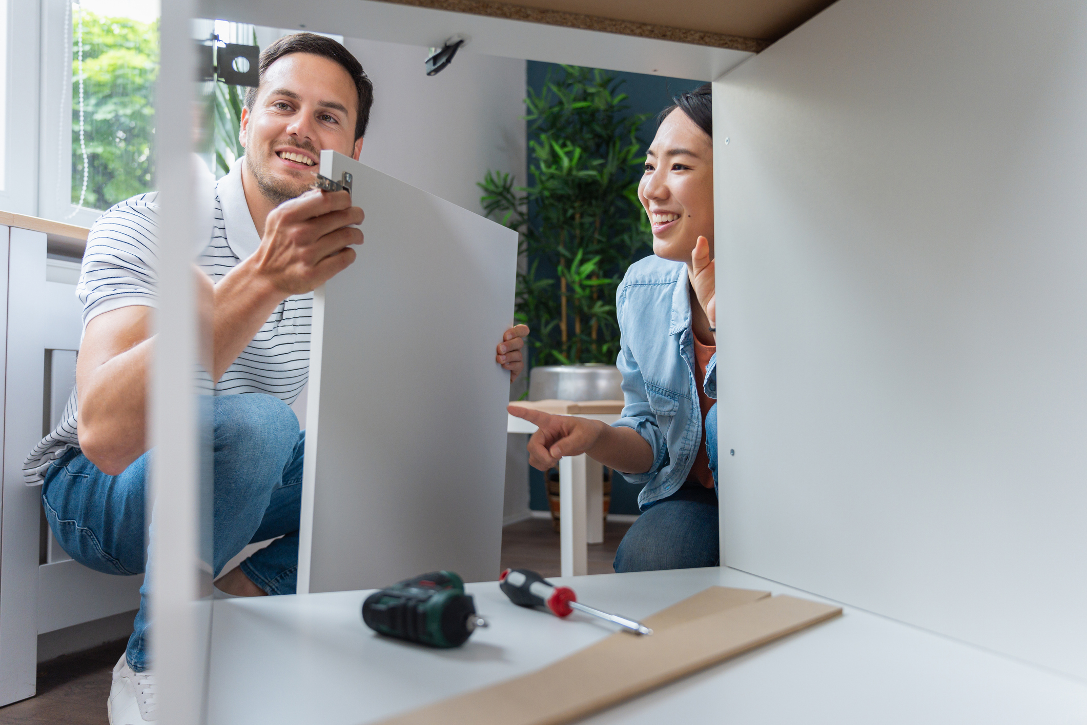 Multiracial couple, assembling white kitchen cabinets in their home.