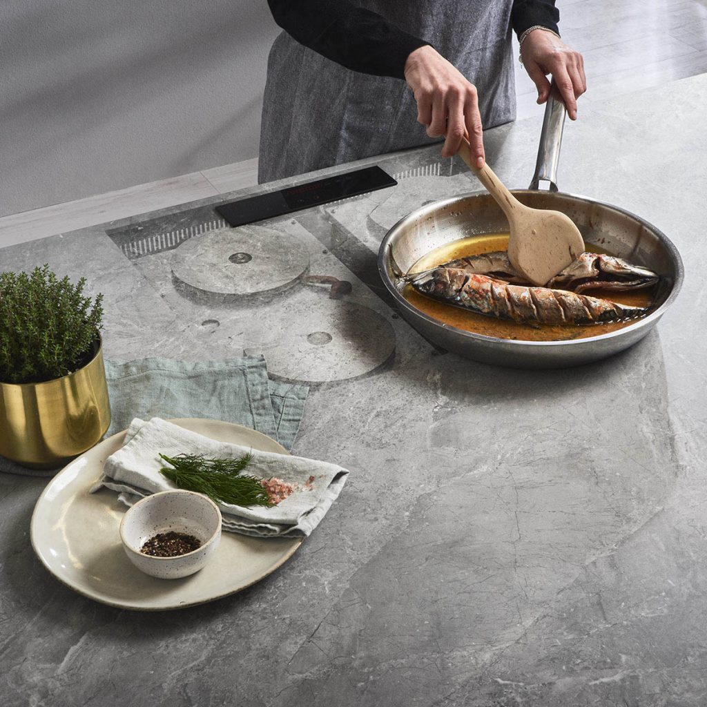 Infinity Invisicook cooktop with faceless woman cooking fish in a pan on top of a gray counter