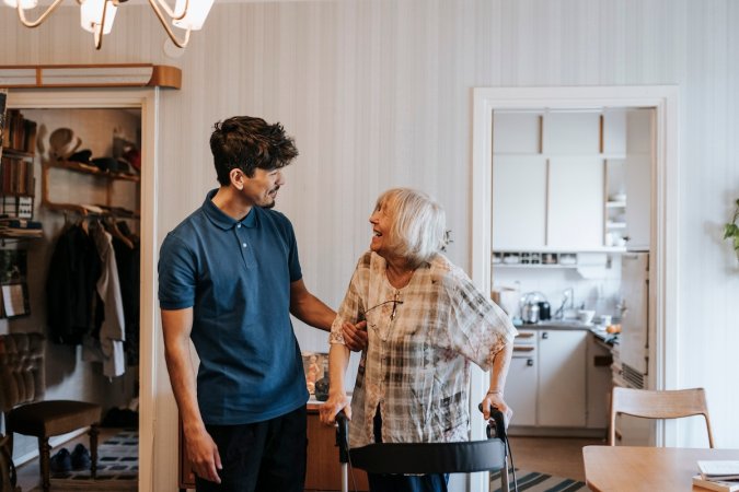 An elderly woman smiles at a young man helping her in her home.