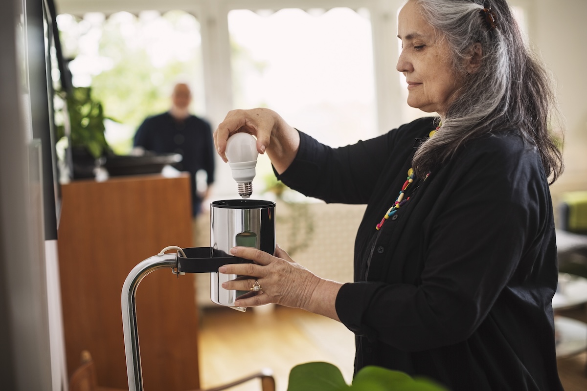 An elderly woman changes a light bulb in a lamp.