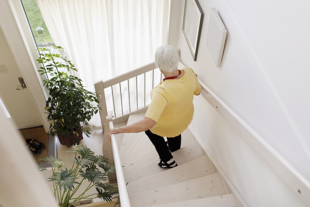 An older woman walks down a staircase with the help of hand railings.