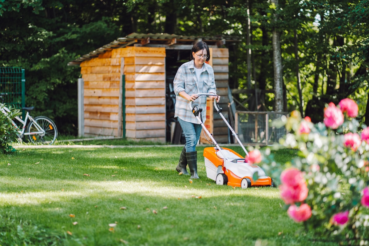 A woman wearing rain boots pushes a lawn mower in her backyard.