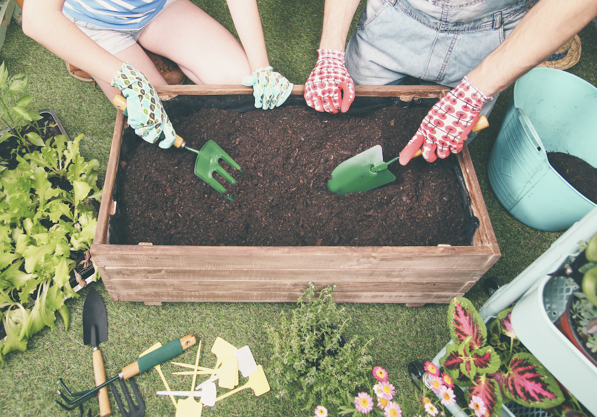Young couple (heads not seen) use trowels to dig into a small raised garden bed on Astroturf, surrounded by gardening accessories.