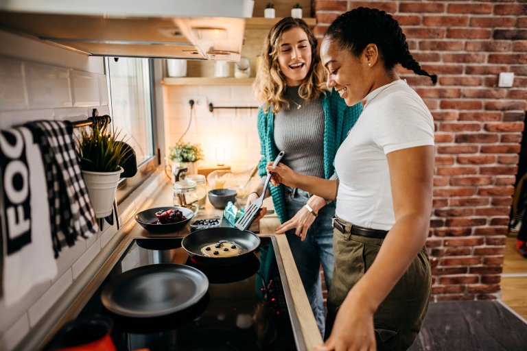 Two women cooking food in a pan on a hot stove in a kitchen.