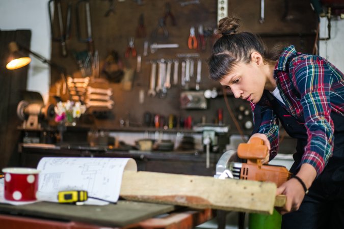 Young woman using a saw in a garage with tools on the wall in the background.