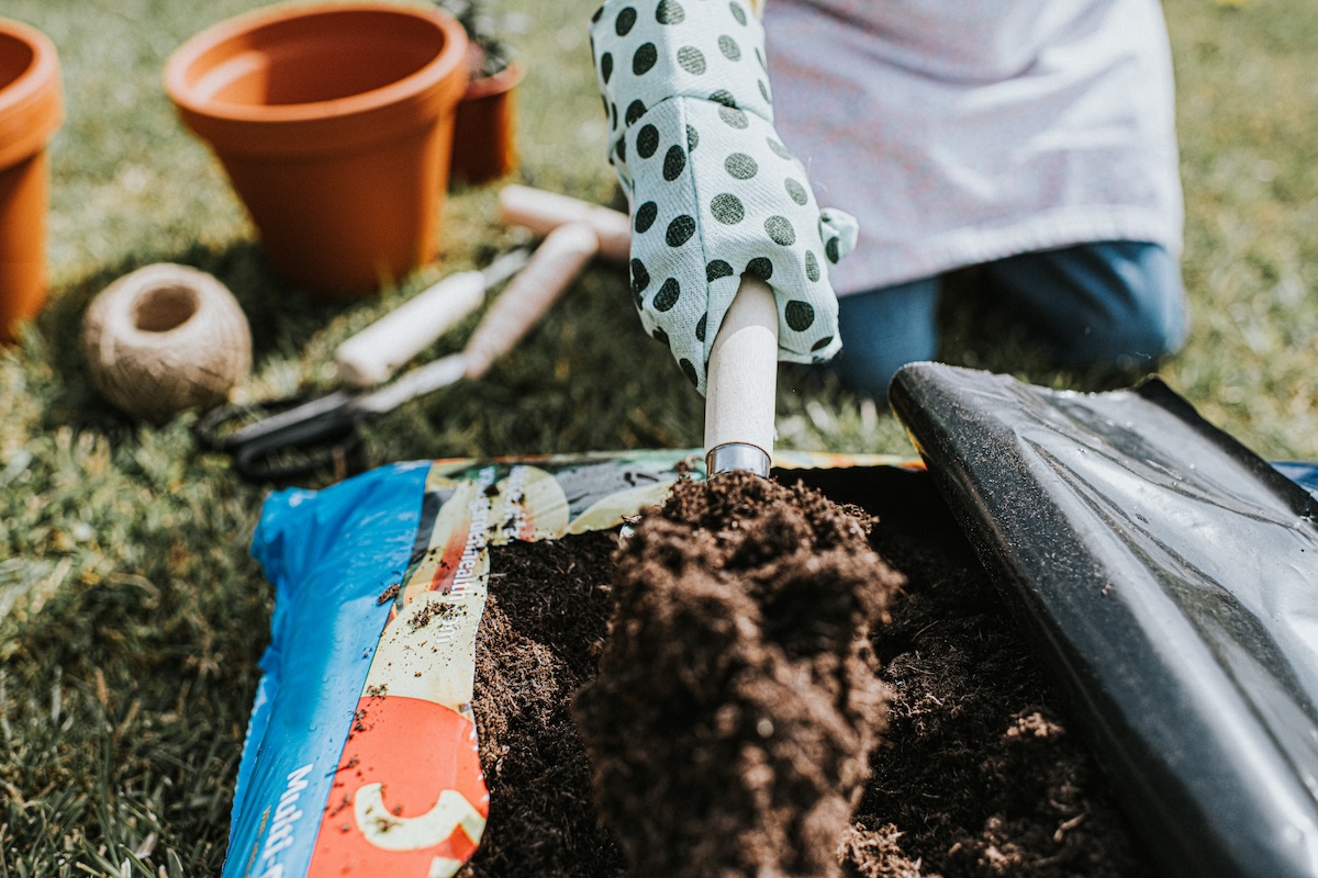 Close-up image of a person kneeling beside a bag of compost and lifting a large scoop of soil out of the bag on a trowel.