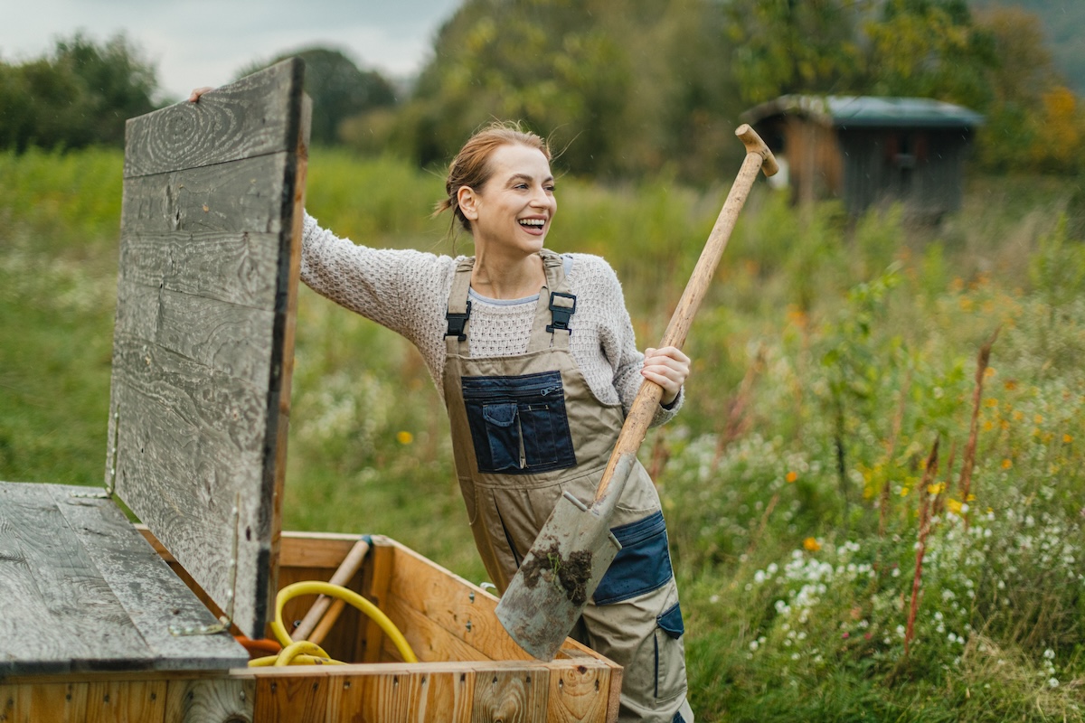A smiling woman with red hair and overalls uses a shovel while gardening.
