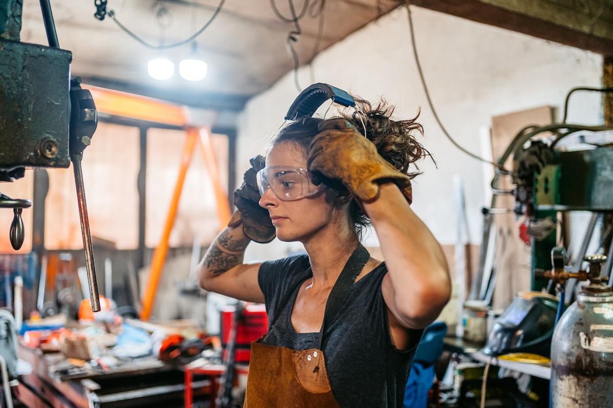 A young woman puts on ear protection, gloves, and eye goggles while working in maintenance shop.