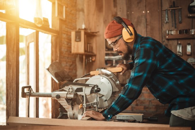 A man wearing personal protective equipment uses a table saw in his home workshop.