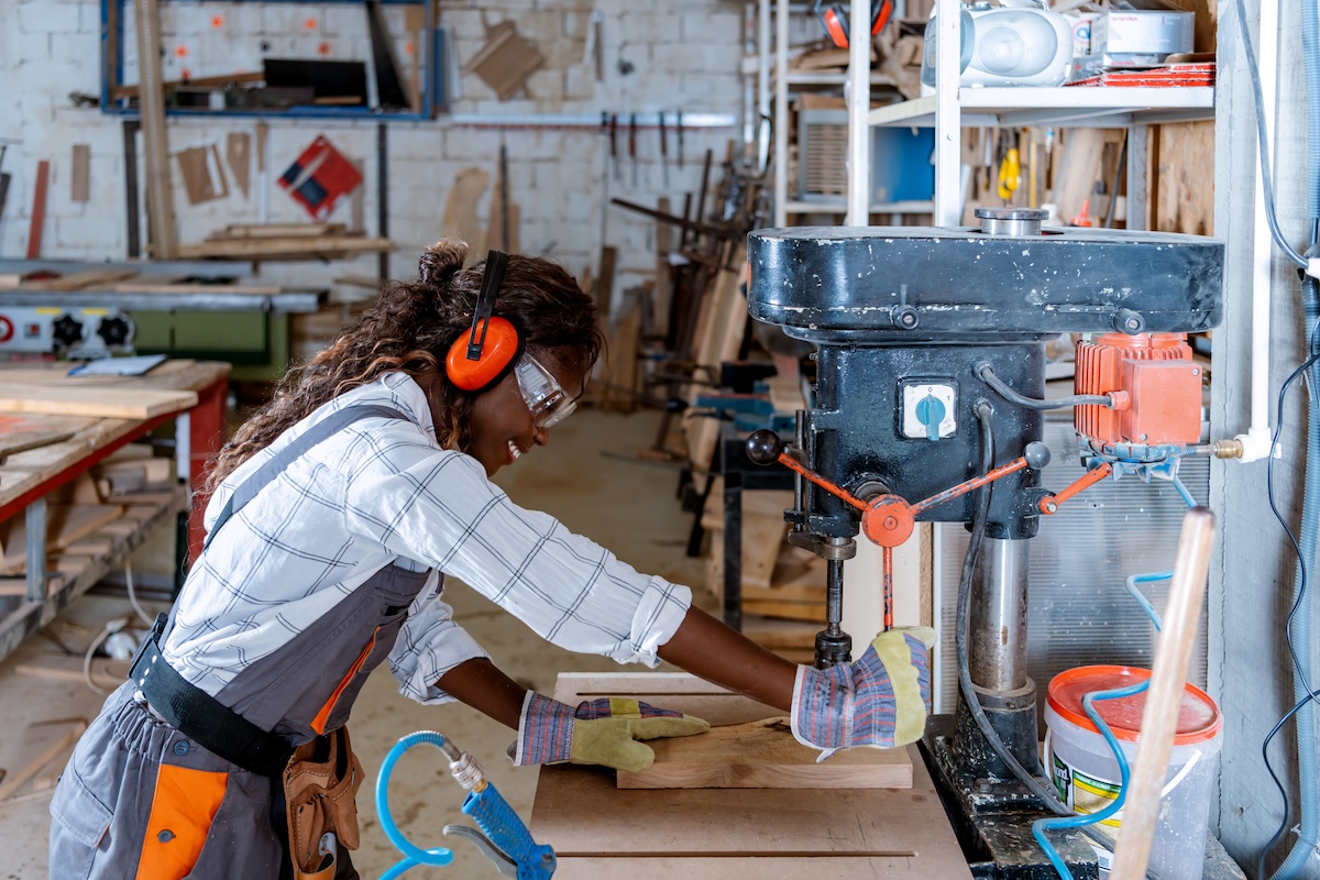 A young carpenter uses a drill press while wearing PPE.