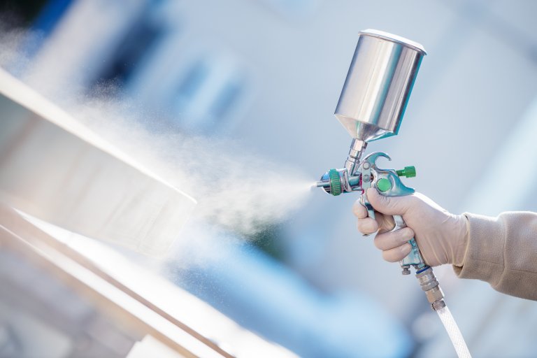 Hand holding a spray gun and painting a piece of wood.