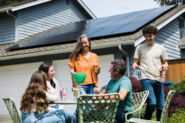 A family with three children having fun connecting with one another outside on their lawn, sitting at an outdoor table. Their solar panel array is visible on the roof of their home.