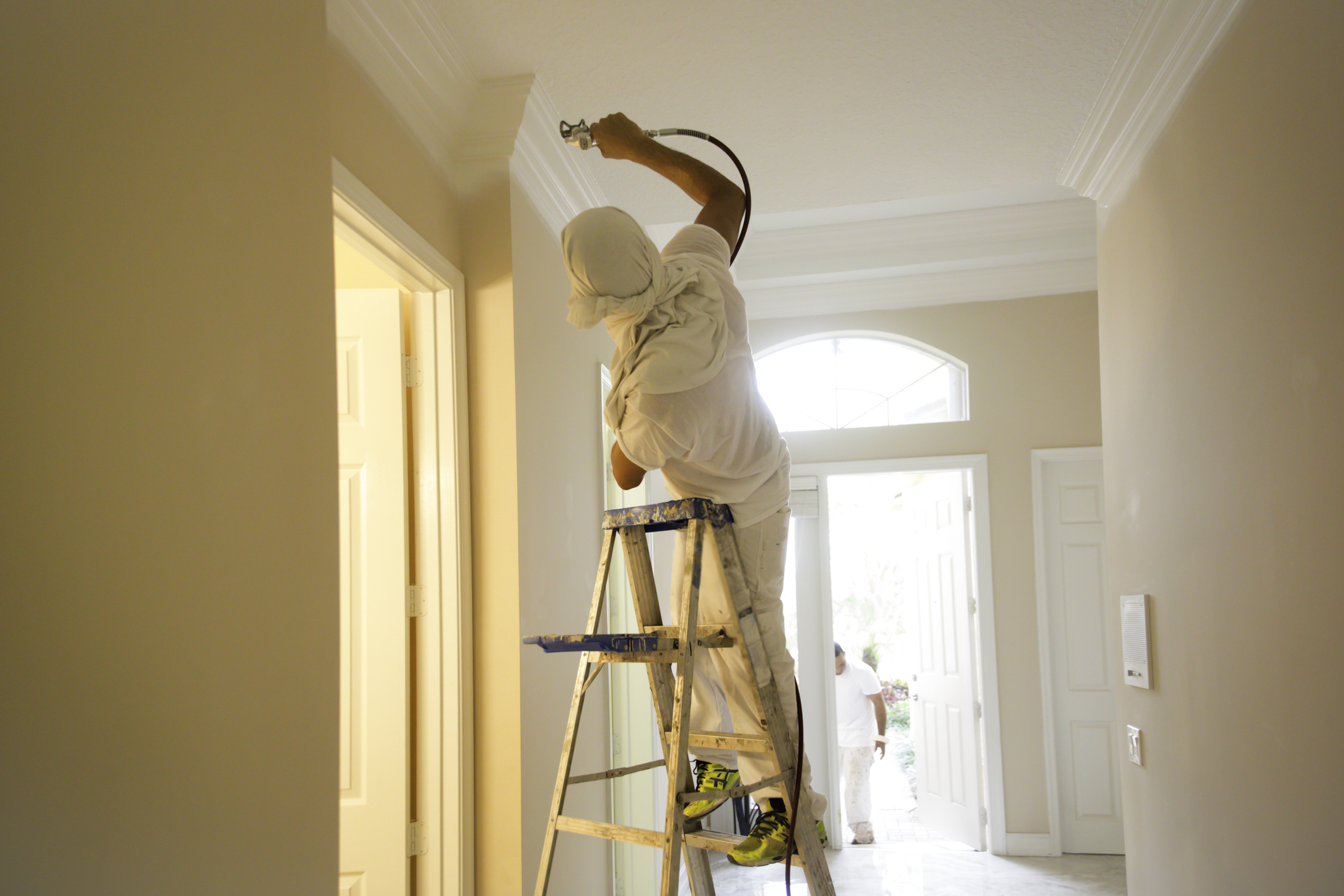 Man using spray paint gun to paint the crown molding on the ceiling in home.