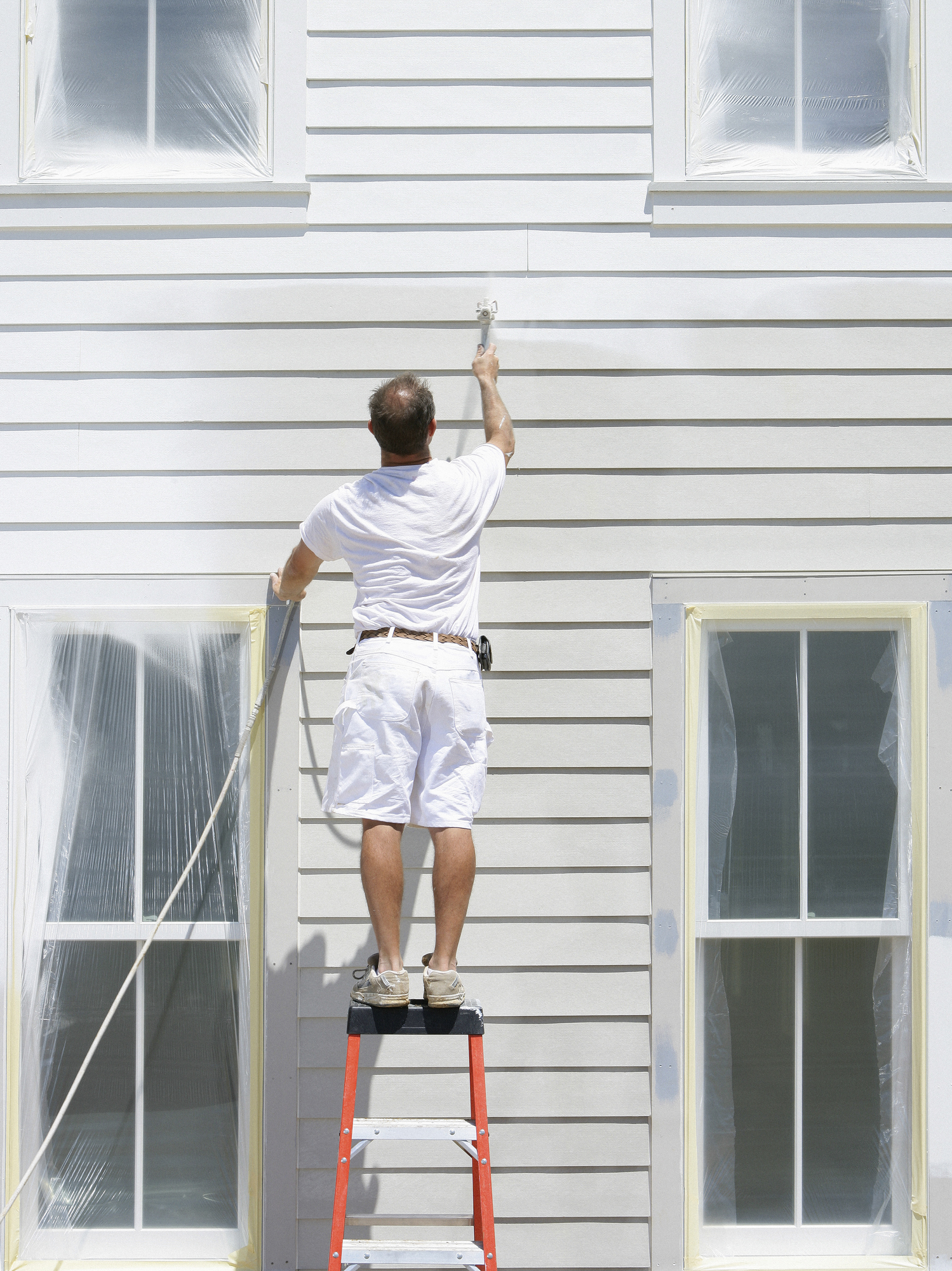 Man standing on a ladder painting exterior of a home with a paint sprayer.