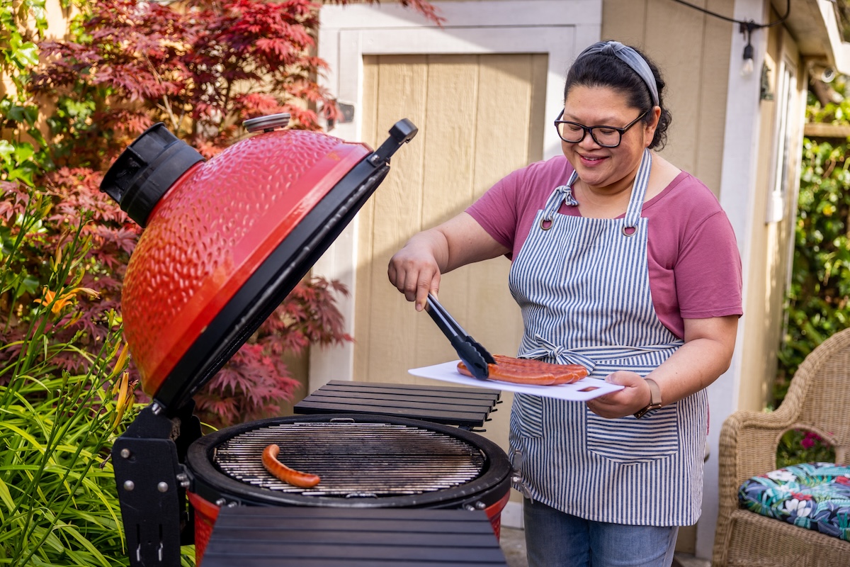 A woman smiles as she grills hotdogs on a red charcoal grill.