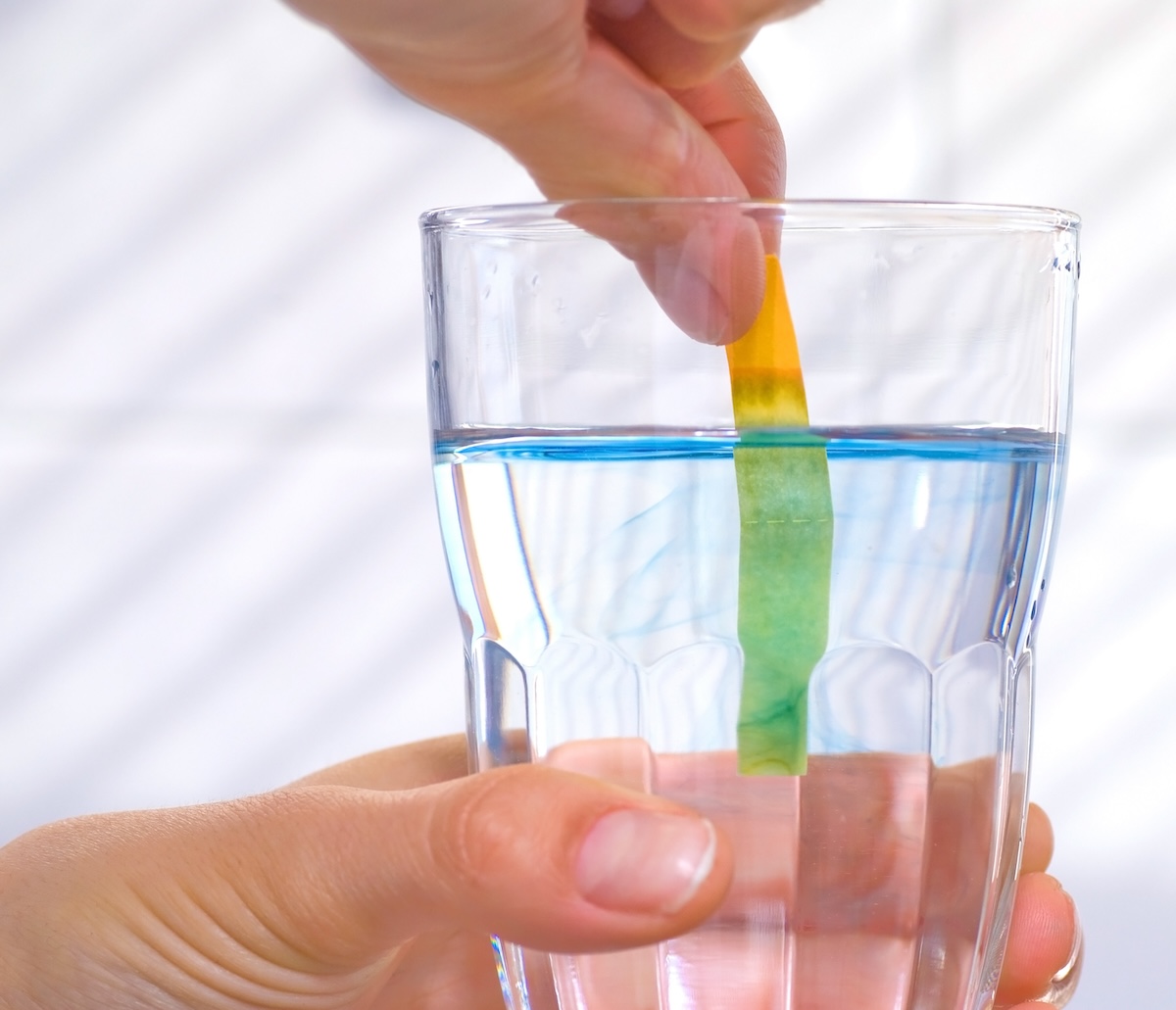 Hands hold a glass of water and dips a testing strip to determine water hardness.