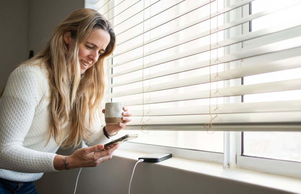 Smiling woman drinking coffee and charging her phone on a windowsill.