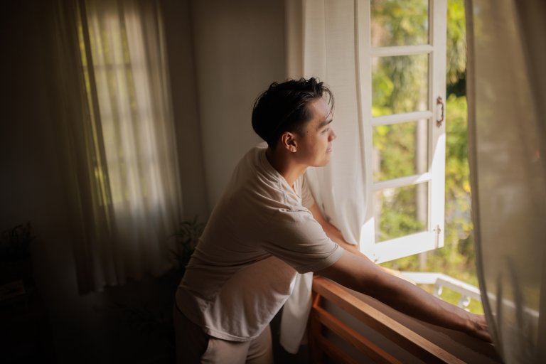 A smiling man looks through an open window after waking up in the morning.