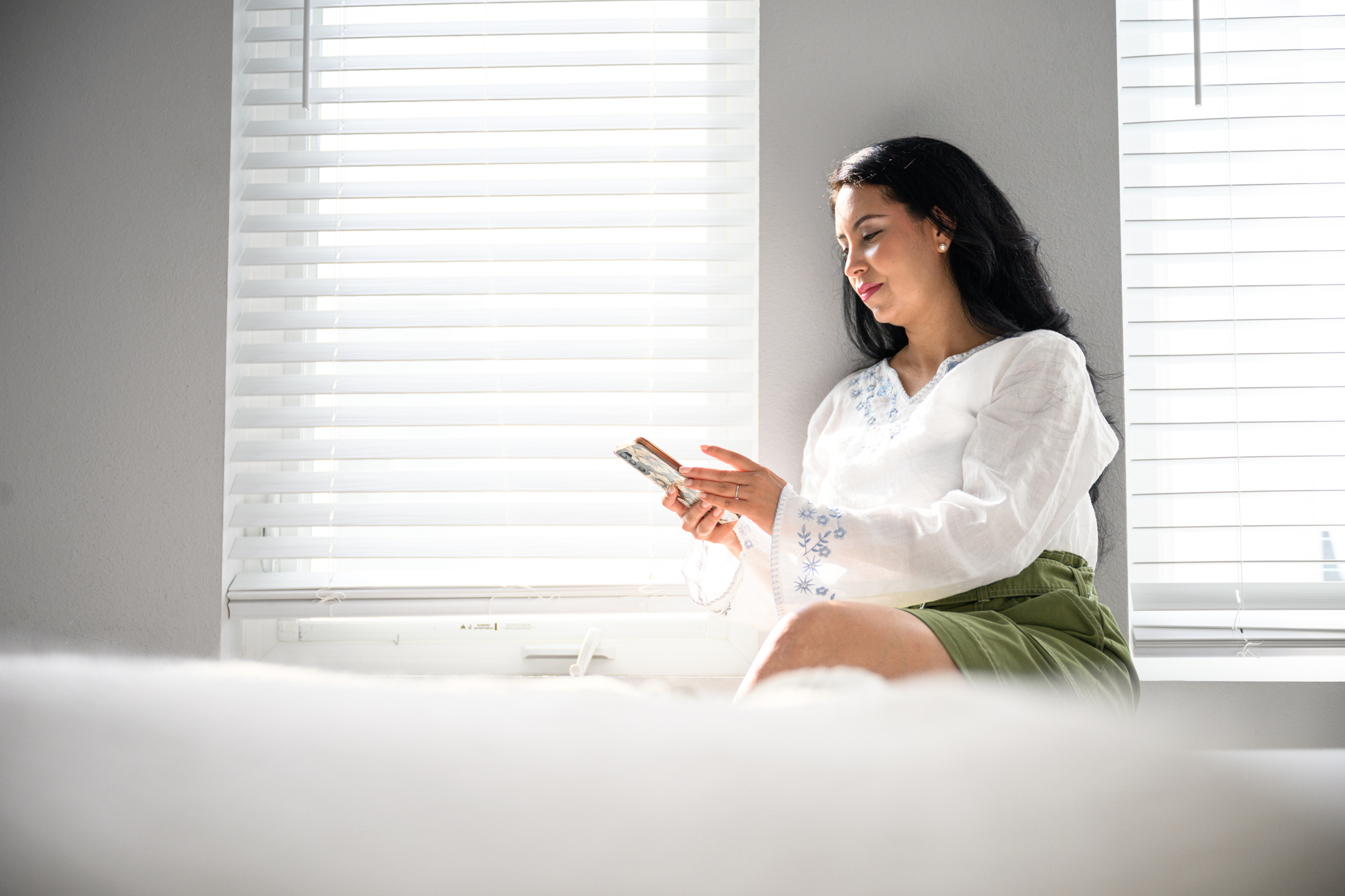 Woman on mobile phone sitting in front of bedroom window with cellular shades