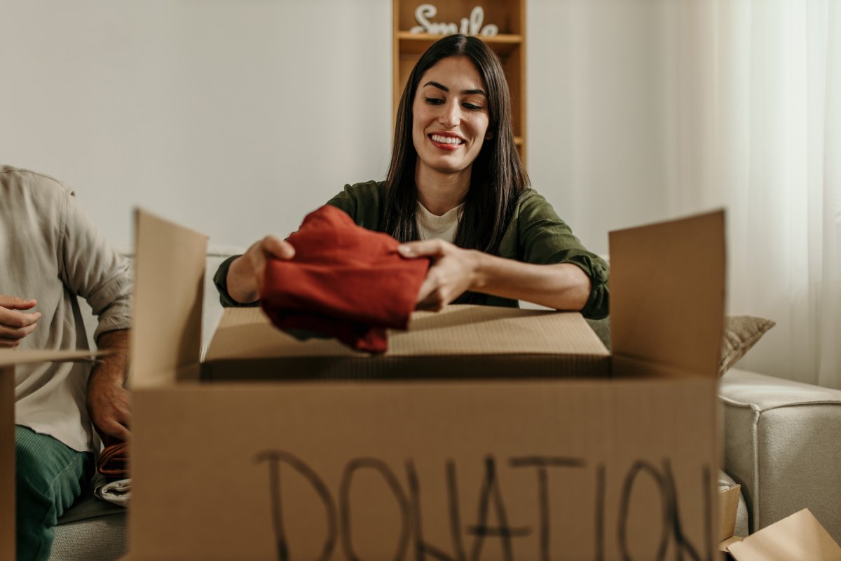 A woman is sitting on a couch putting clothing into a cardboard box labeled donation