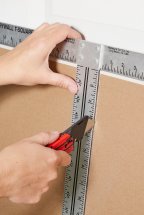 Hands using a utility knife to score a sheet of drywall.