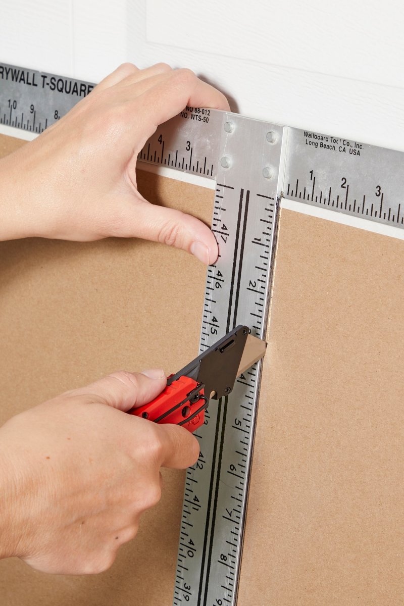 Hands using a utility knife to score a sheet of drywall.