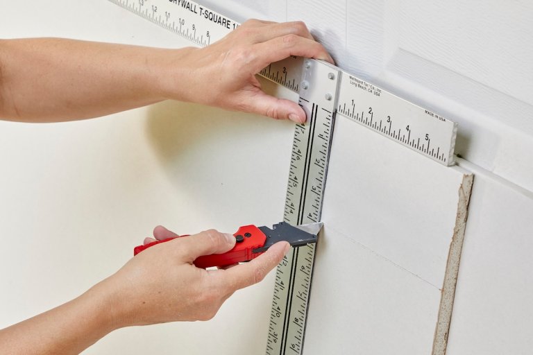 A person cutting a piece of drywall with a utility knife and a T-square.