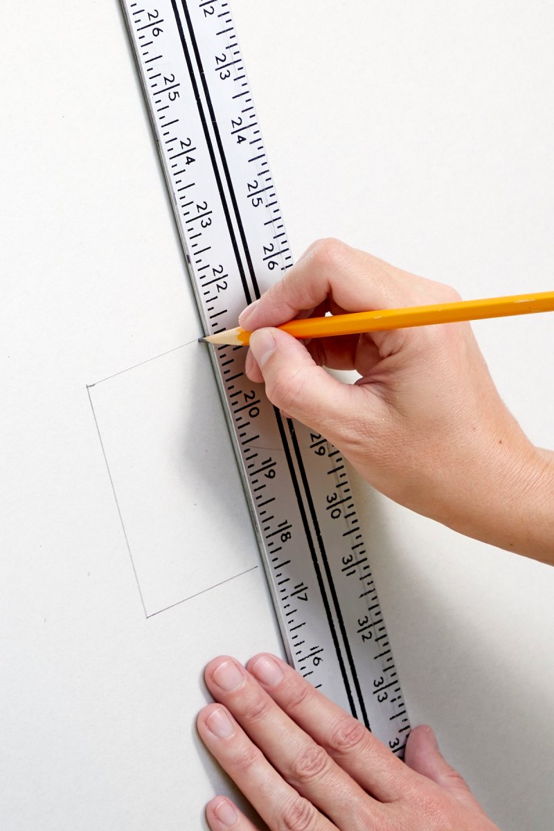 Hands using a pencil and T-square to mark cut location on sheet of drywall.
