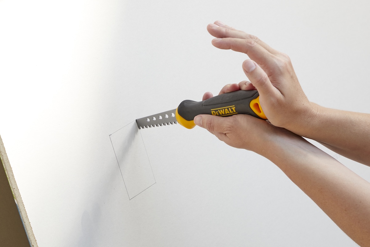 Hands using a jab saw to cut a hole in drywall for an electrical outlet.